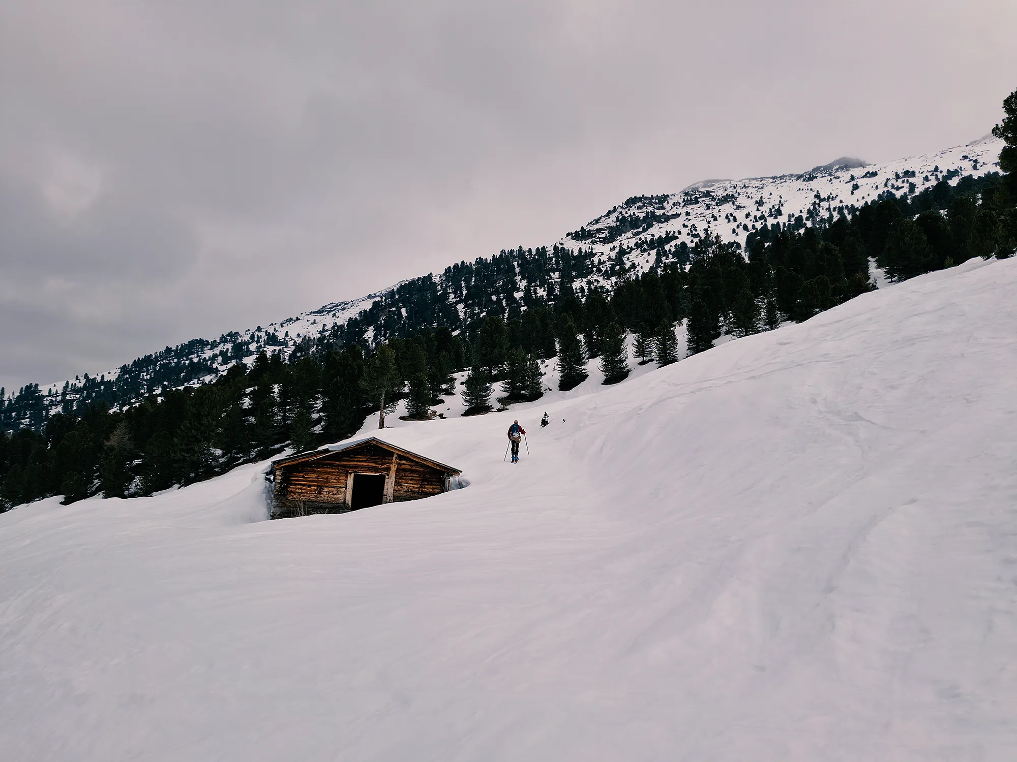 Verschneite Berglandschaft. Ein Skitourengeher geht an einer kleinen Holzhütte vorbei.